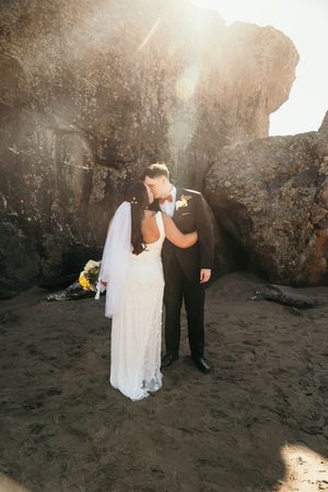 Bride and groom share a romantic kiss on a sandy beach surrounded by dramatic rocks with golden sunlight streaming behind them.