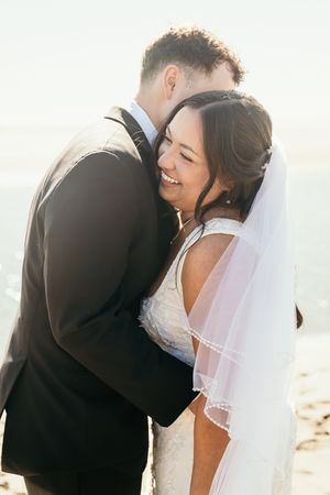 Bride smiling joyfully as groom embraces her on a sunlit beach in wedding attire.