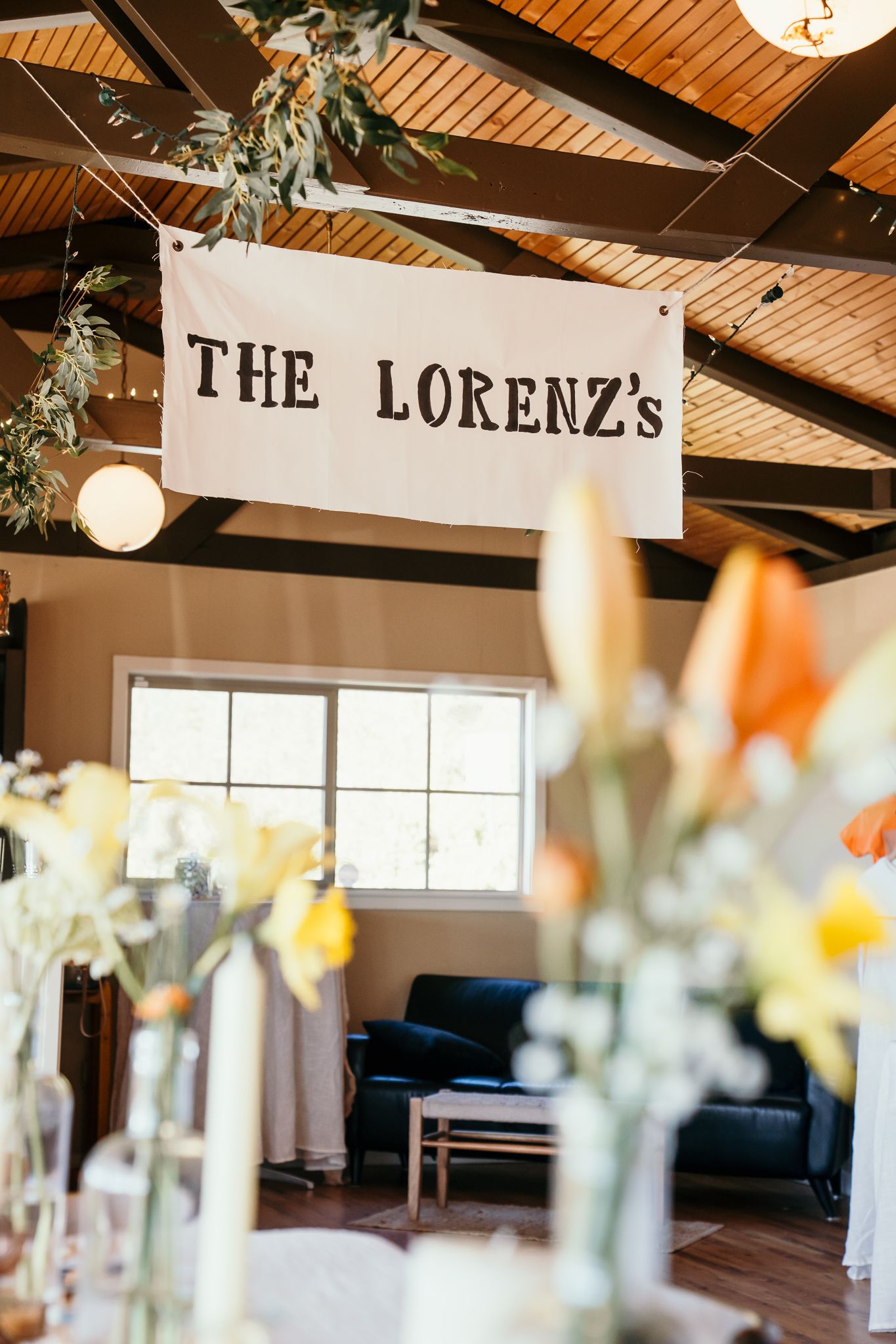A rustic wedding reception venue decorated with a hanging banner reading The Lorenzs, floral arrangements, and string lights.