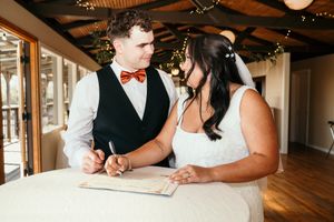 A bride and groom smile while signing their marriage certificate at a rustic indoor wedding venue.