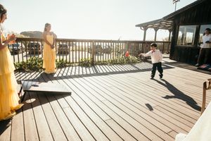 Child playing cornhole on a sunny wooden deck at an outdoor venue with adults in yellow dresses watching.