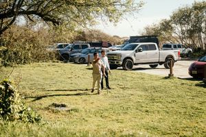 Two people standing on a grassy area near a parking lot with trucks and cars on a sunny day.