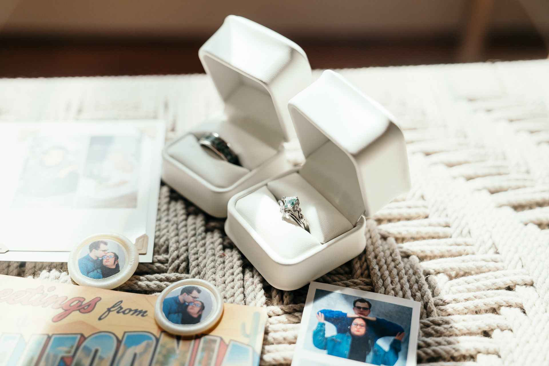 Two silver rings displayed in a white jewelry box surrounded by photo prints and keepsakes on a woven surface.