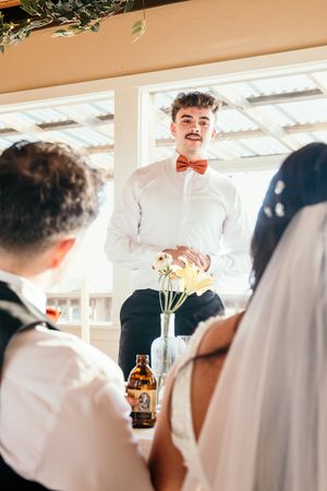 Young man in white shirt and rust bow tie giving a wedding speech to seated bride and groom at reception.