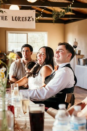 Bride and groom laugh joyfully at their wedding reception under a "The Lorenz's" banner with guests seated at decorated tables.