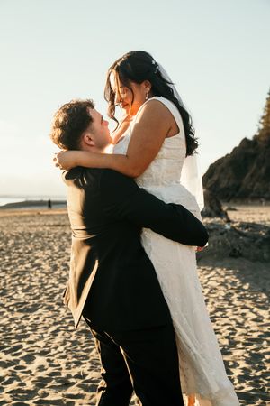 Groom lifts smiling bride in white dress on a sunlit beach with rocky cliffs in the background during golden hour.