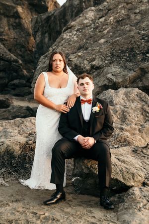 Bride in white lace gown and groom in black tuxedo with rust bow tie posing on rocks outdoors.