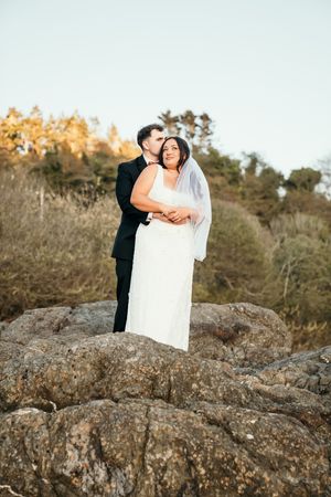 Bride and groom standing on rocky terrain, embracing outdoors with autumn trees in the background.