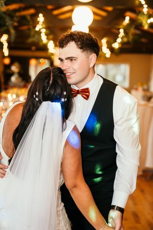 Groom in black vest and red bow tie shares first dance with bride in veil under warm string lights at reception.