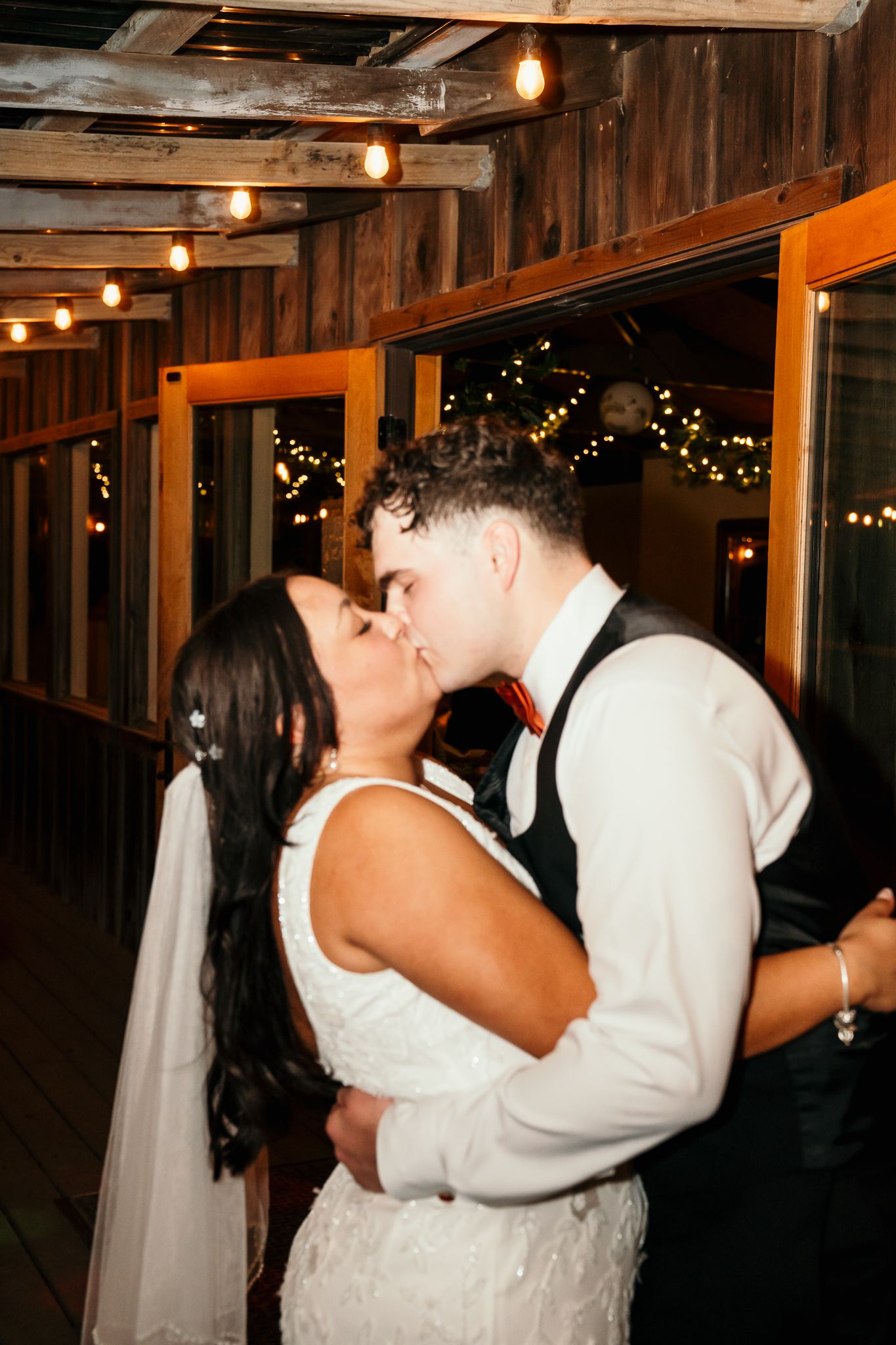 A bride and groom share a romantic kiss at their wedding reception, surrounded by warm string lights in a rustic venue.