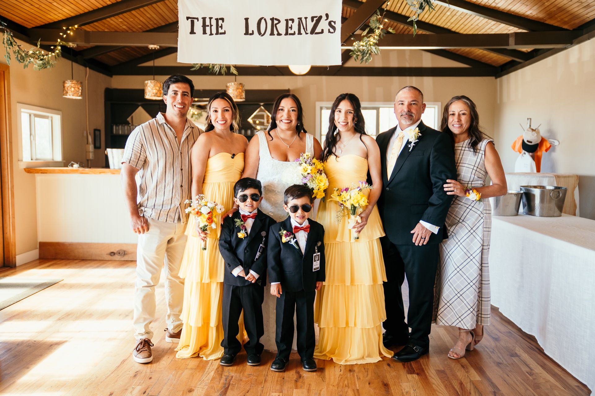 Wedding party in yellow bridesmaid dresses with two ring bearers in suits posing indoors under The Lorenzs sign.