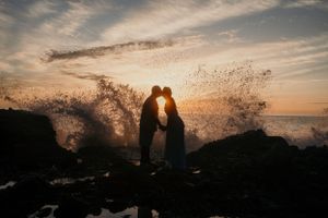 Silhouetted couple shares intimate moment as ocean wave crashes behind them during sunset.