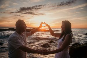 Two silhouettes make a heart shape with their hands against a vibrant ocean sunset backdrop.