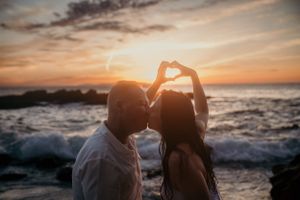 Silhouetted couple making heart shape with hands against dramatic ocean sunset.