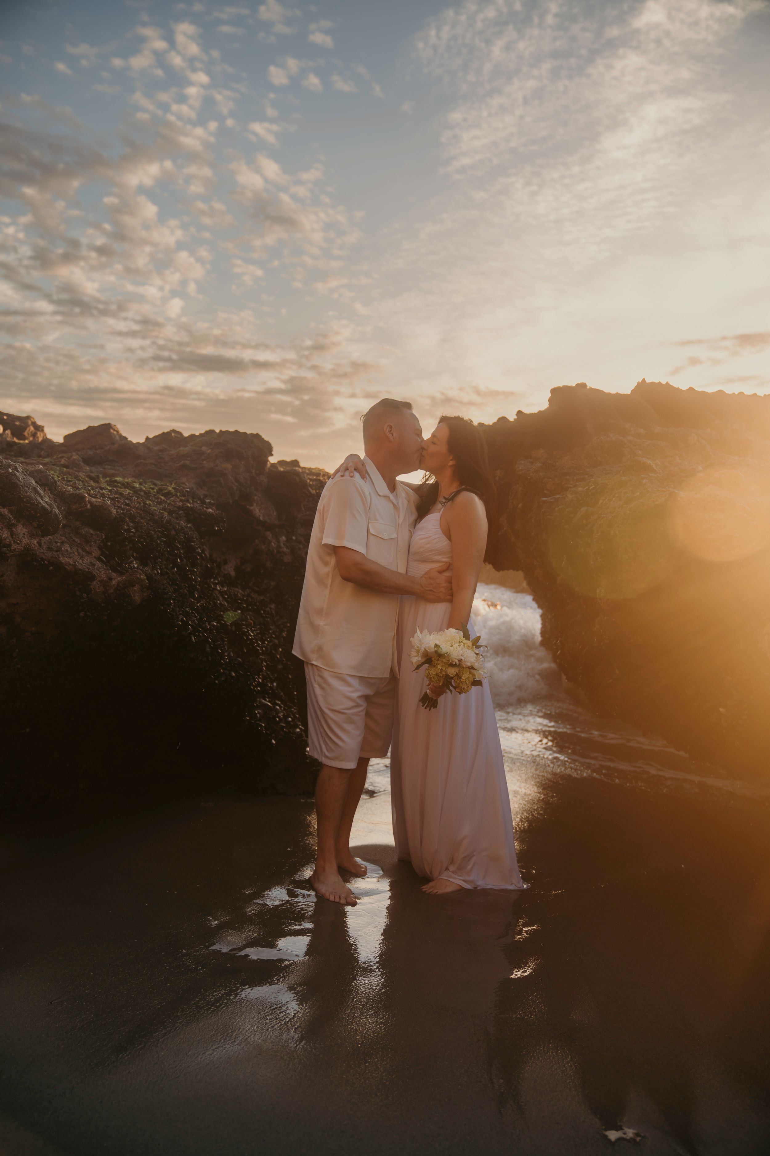 A couple shares a romantic moment on wet volcanic rocks during a golden sunset by the ocean.