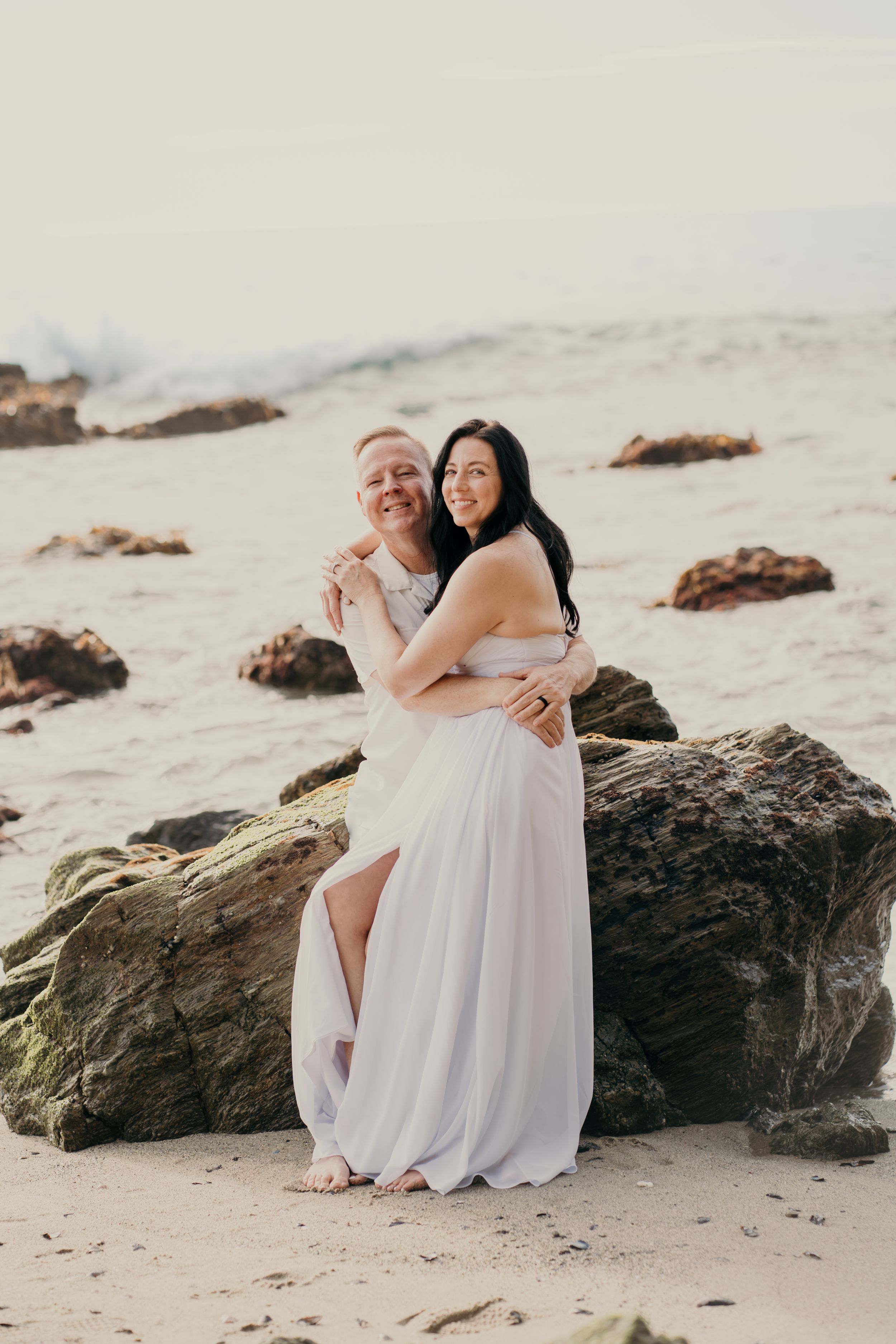 Happy couple in light flowy outfits embrace on a rocky beach during a romantic photoshoot at sunset.