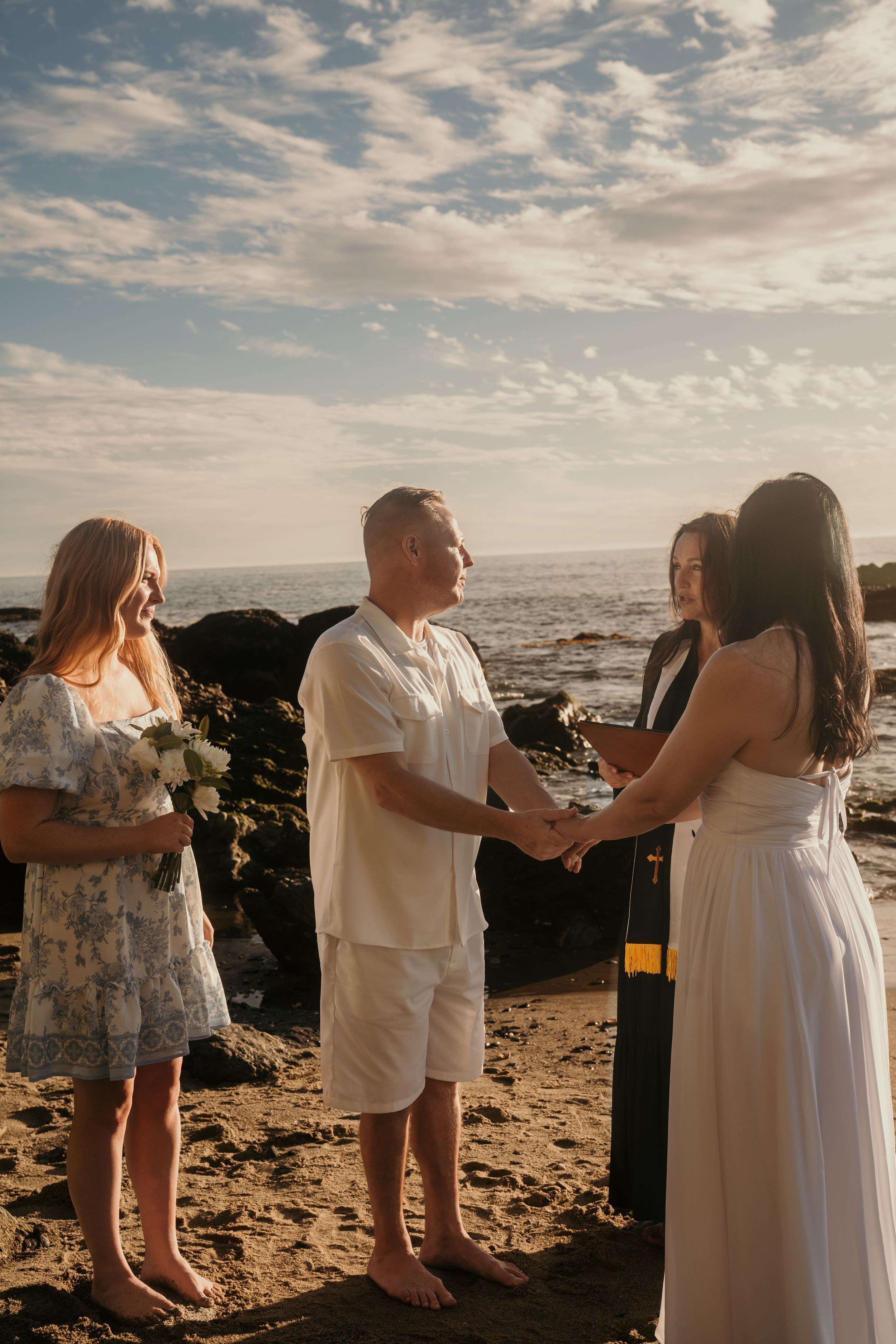 An intimate beach wedding ceremony takes place at sunset with dark rocks and ocean waves in the background.