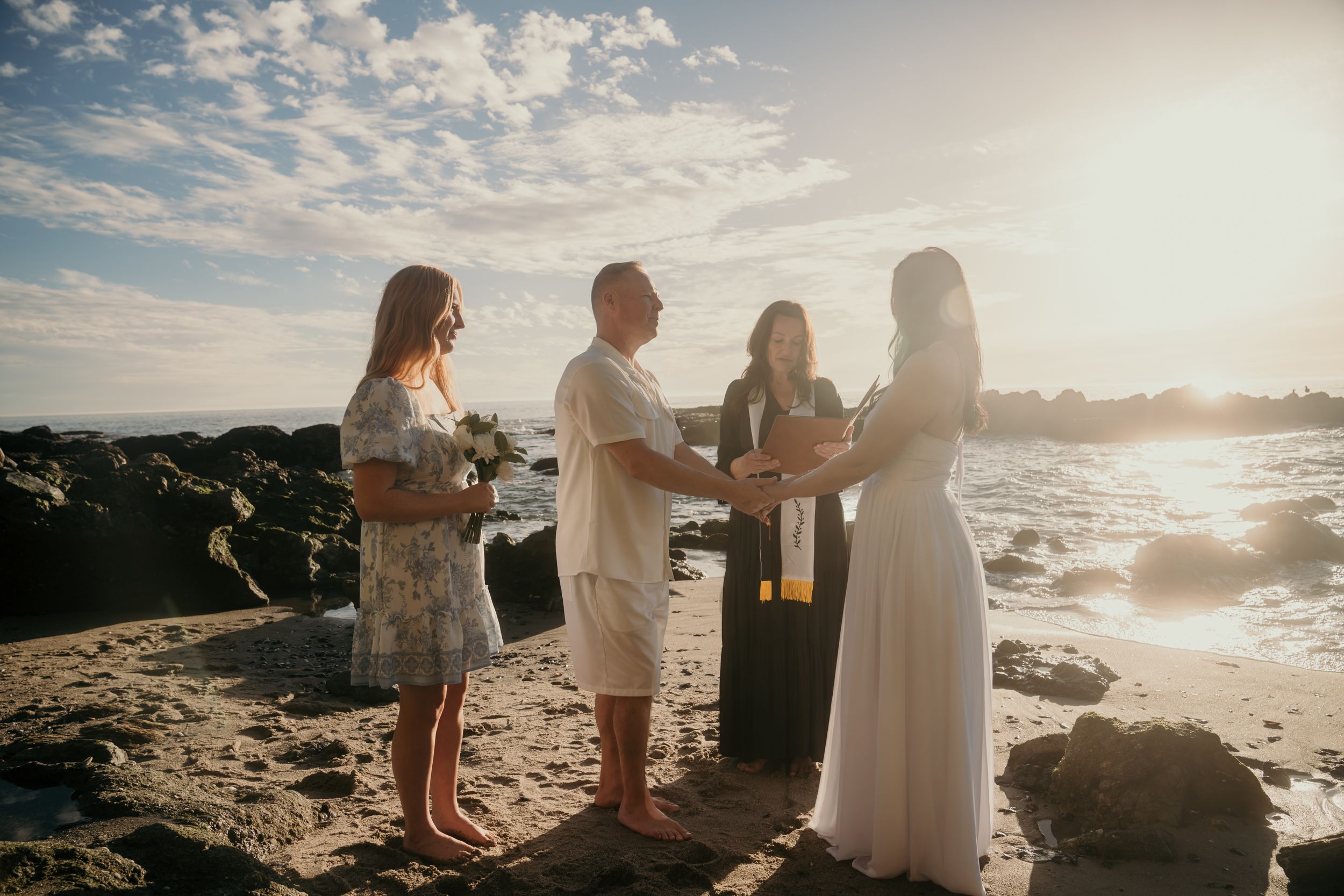Small beachside wedding ceremony occurring at sunset with an officiant and couple on a rocky shore.