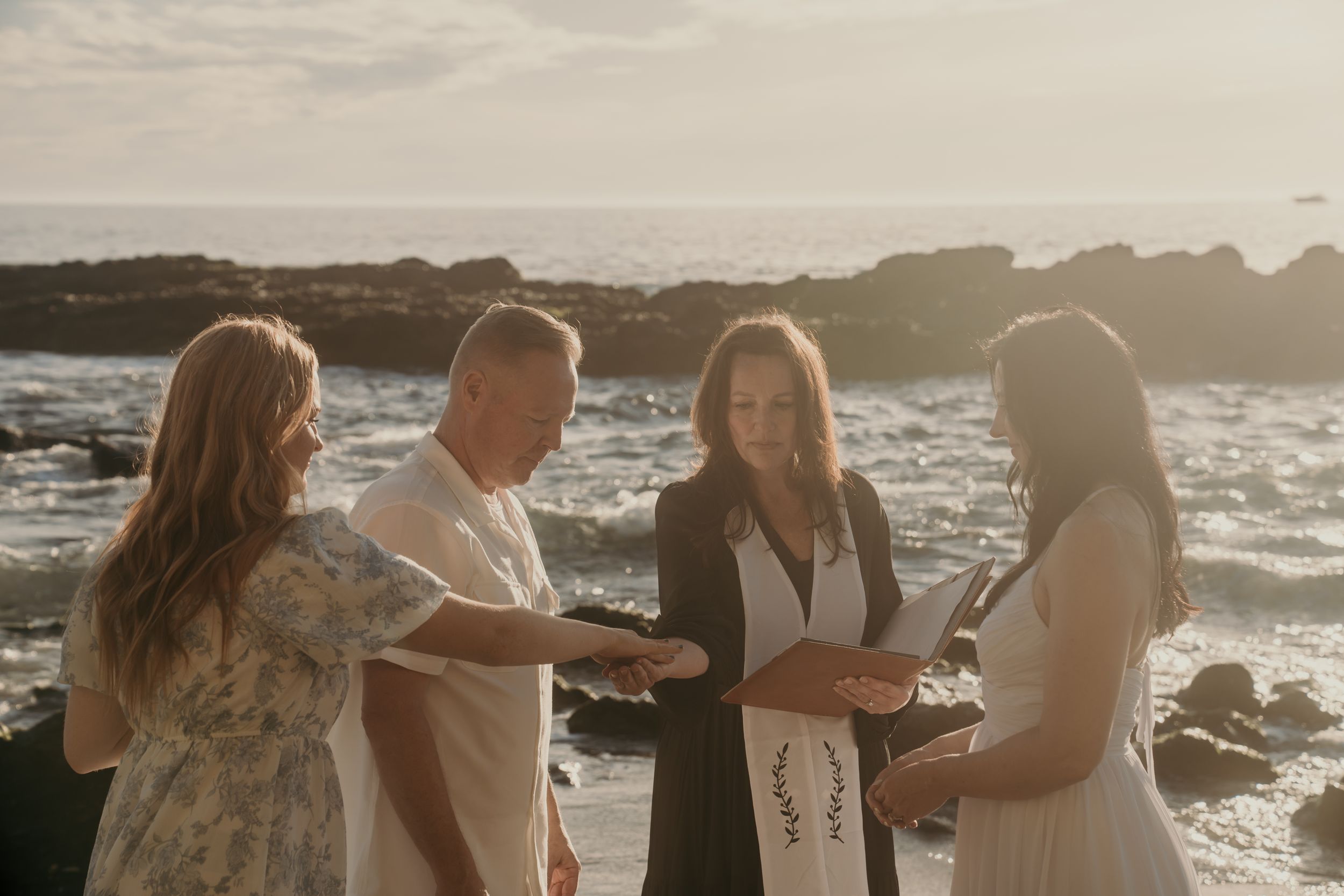 A couple exchanges vows during an intimate beach wedding ceremony at sunset with waves crashing on rocks in the background.