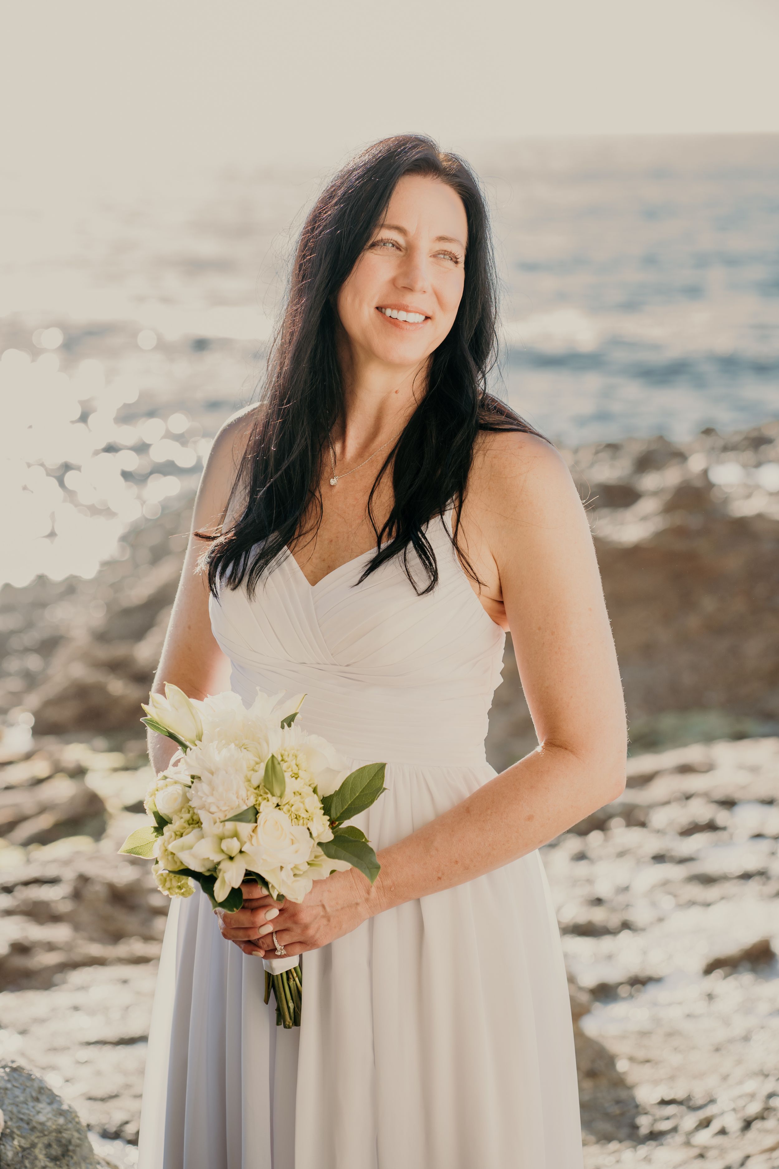 A gorgeous oceanside wedding photo showing someone in a white dress holding a delicate bouquet of cream-colored flowers.