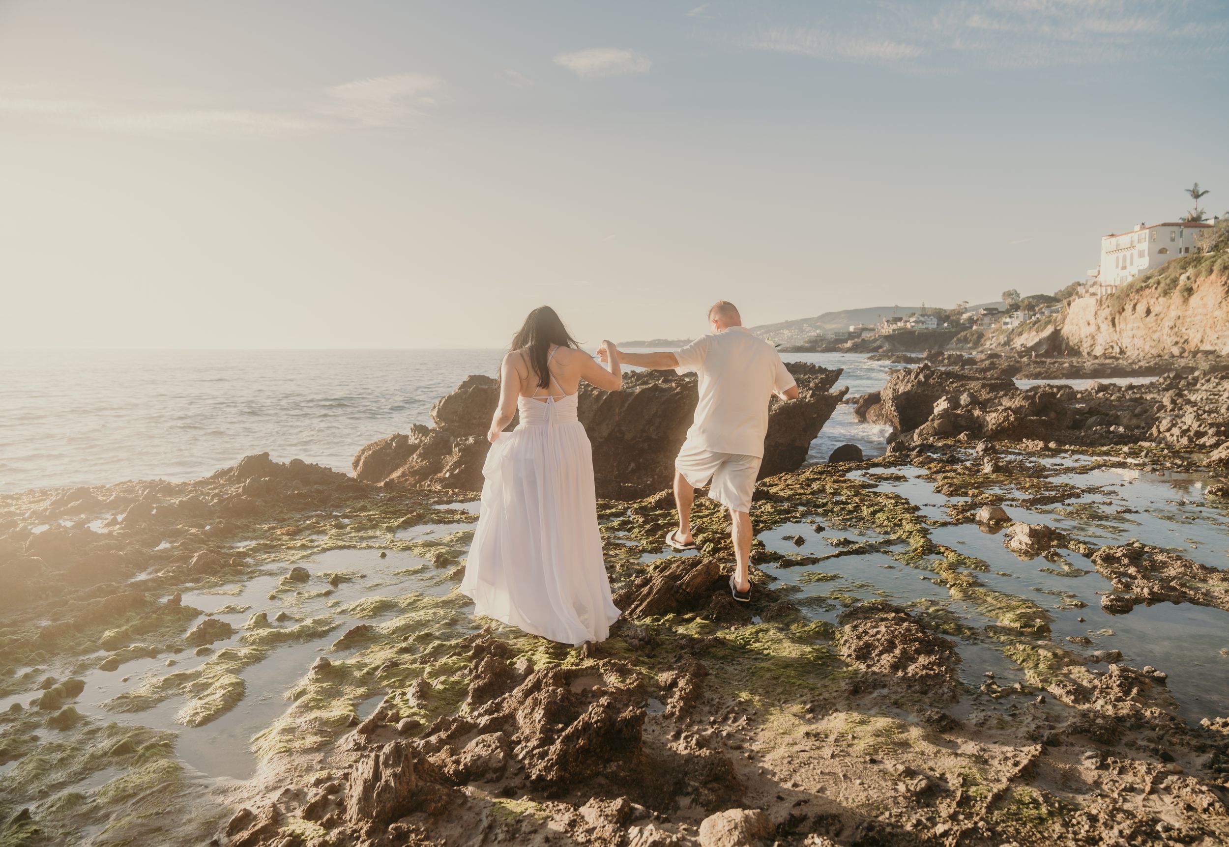 Two people in white dresses stand on coastal rocks at golden hour overlooking the ocean waves.