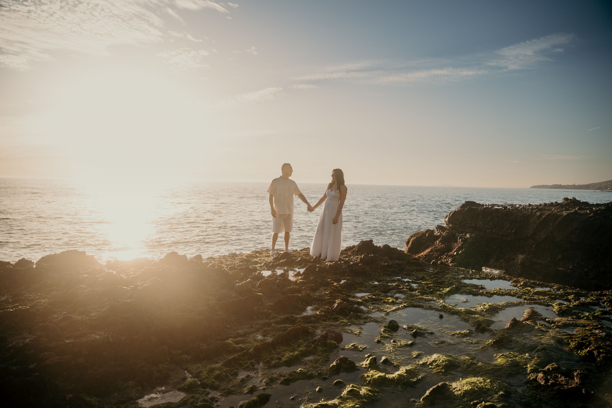 Silhouetted couple holding hands on rocky beach at sunset with ocean waves and glowing horizon in background.