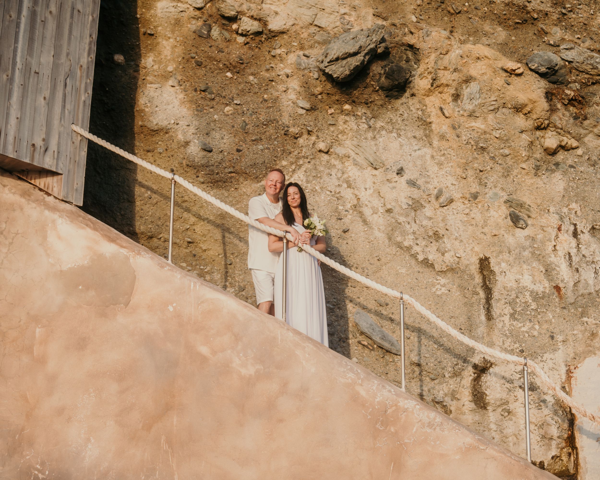 A figure in a white dress stands on a stone stairway against a rustic beige wall with a metal railing.