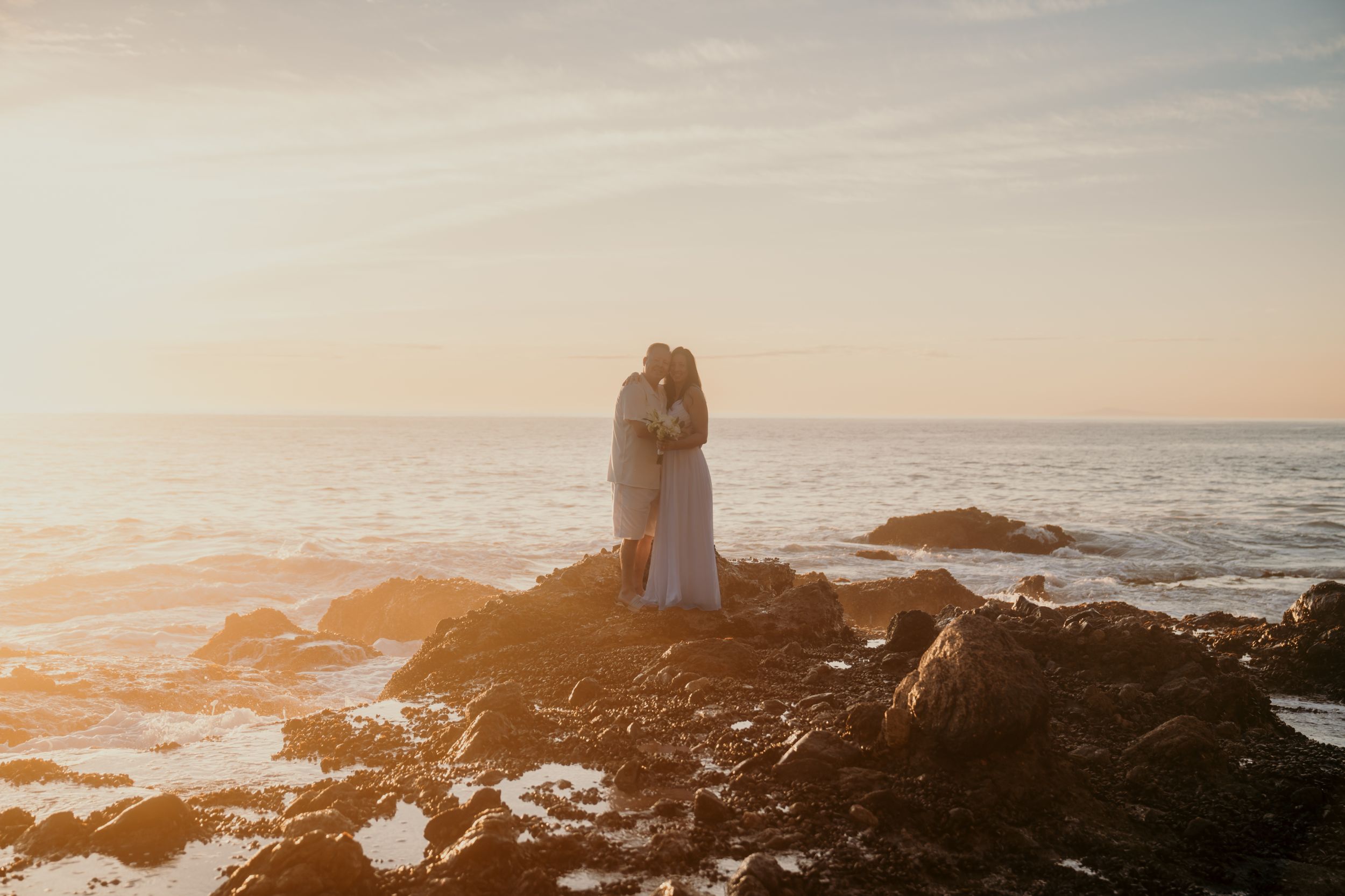 Couple embracing on rocky coastline during dramatic golden sunset over the Pacific ocean.