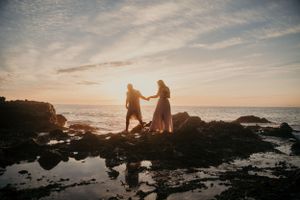 A romantic couple stands silhouetted against a golden sunset on a rocky beach coastline.