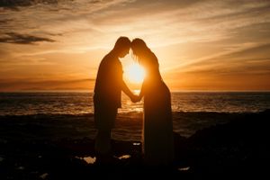 Two silhouettes share an intimate moment against a dramatic orange sunset over the ocean.