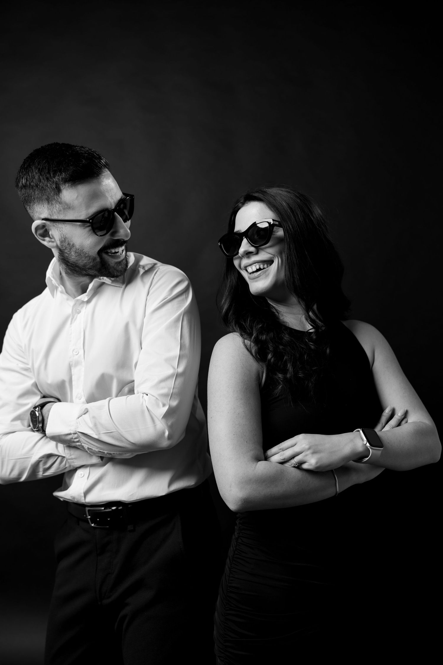 Couple in formal wear sharing a laugh while wearing sunglasses in black and white.