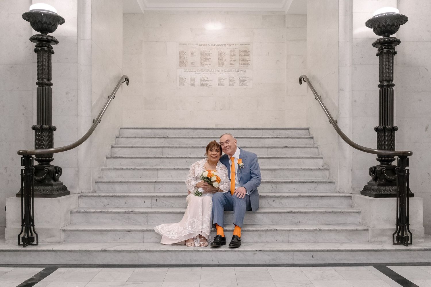 A couple sits together on elegant marble stairs flanked by ornate black lampposts in a light-filled interior space.