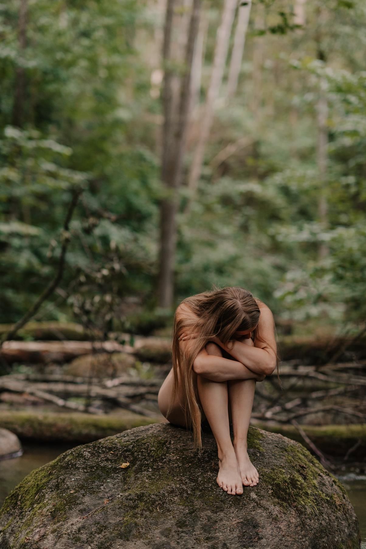 Person in a brown dress curled up on a large mossy boulder in a lush green forest setting.