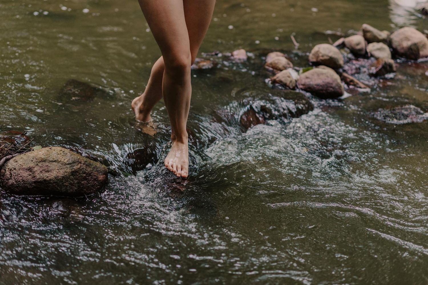 Barefoot legs walking along rocky riverbank splashing in shallow stream water during summer outdoor activity.