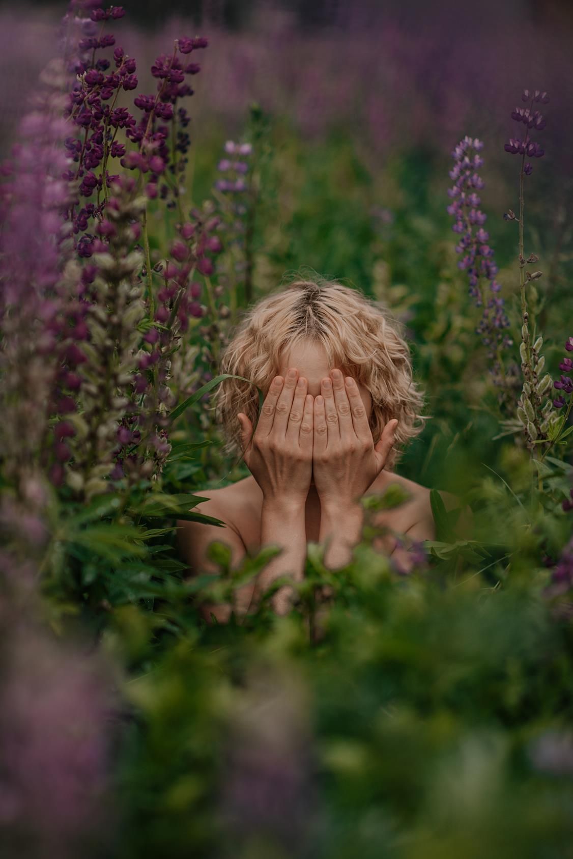 A figure partially hidden among purple flowers and green foliage in a dreamy outdoor garden setting.