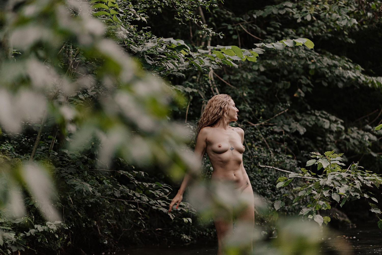 Figure posed among lush green foliage in dappled sunlight.