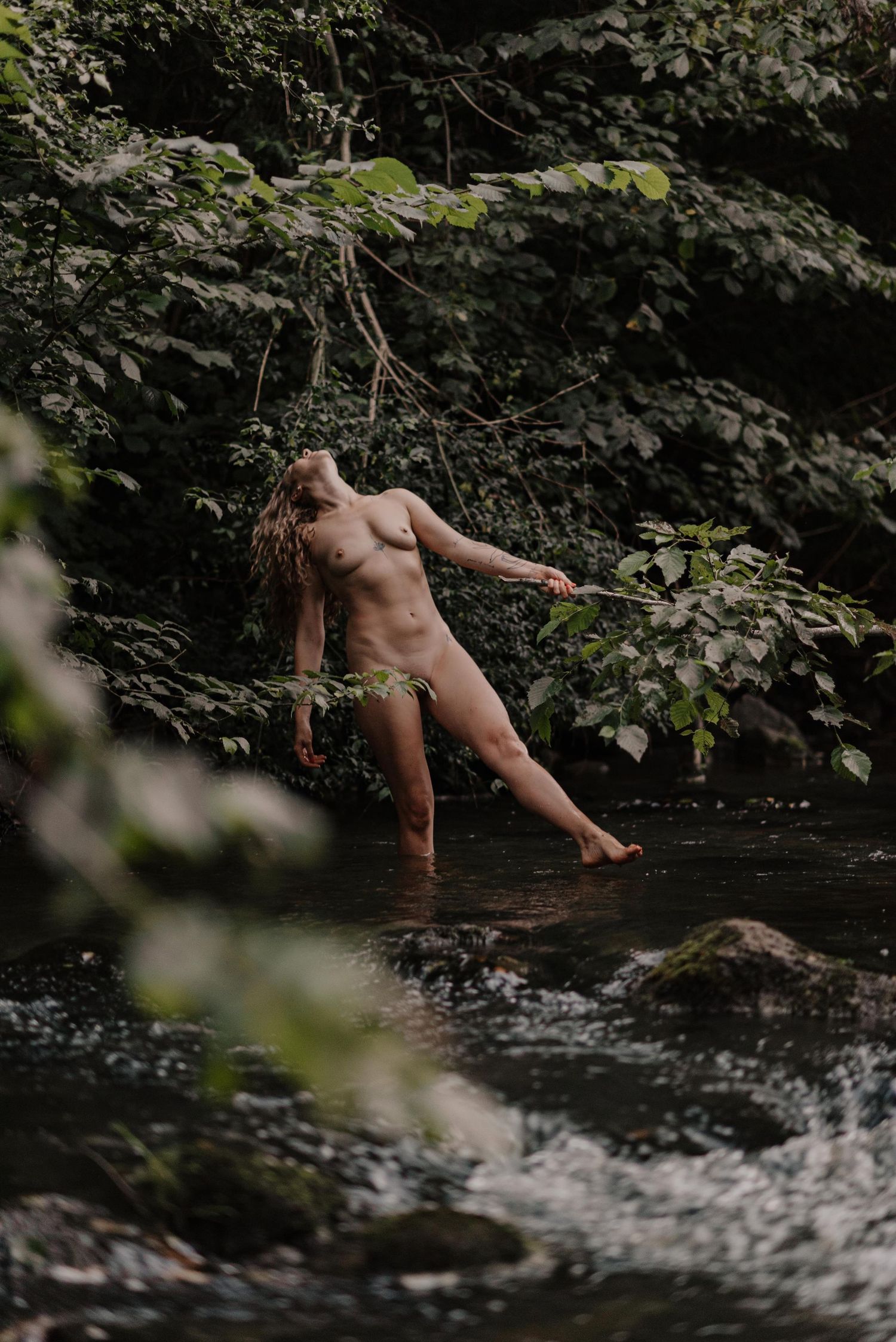 A person splashes and plays in a dark forest stream surrounded by lush greenery.