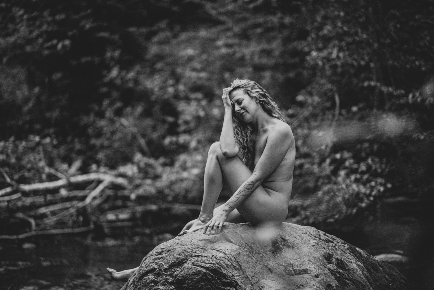 Artistic black and white nude figure poses thoughtfully on a rock by a stream in nature.