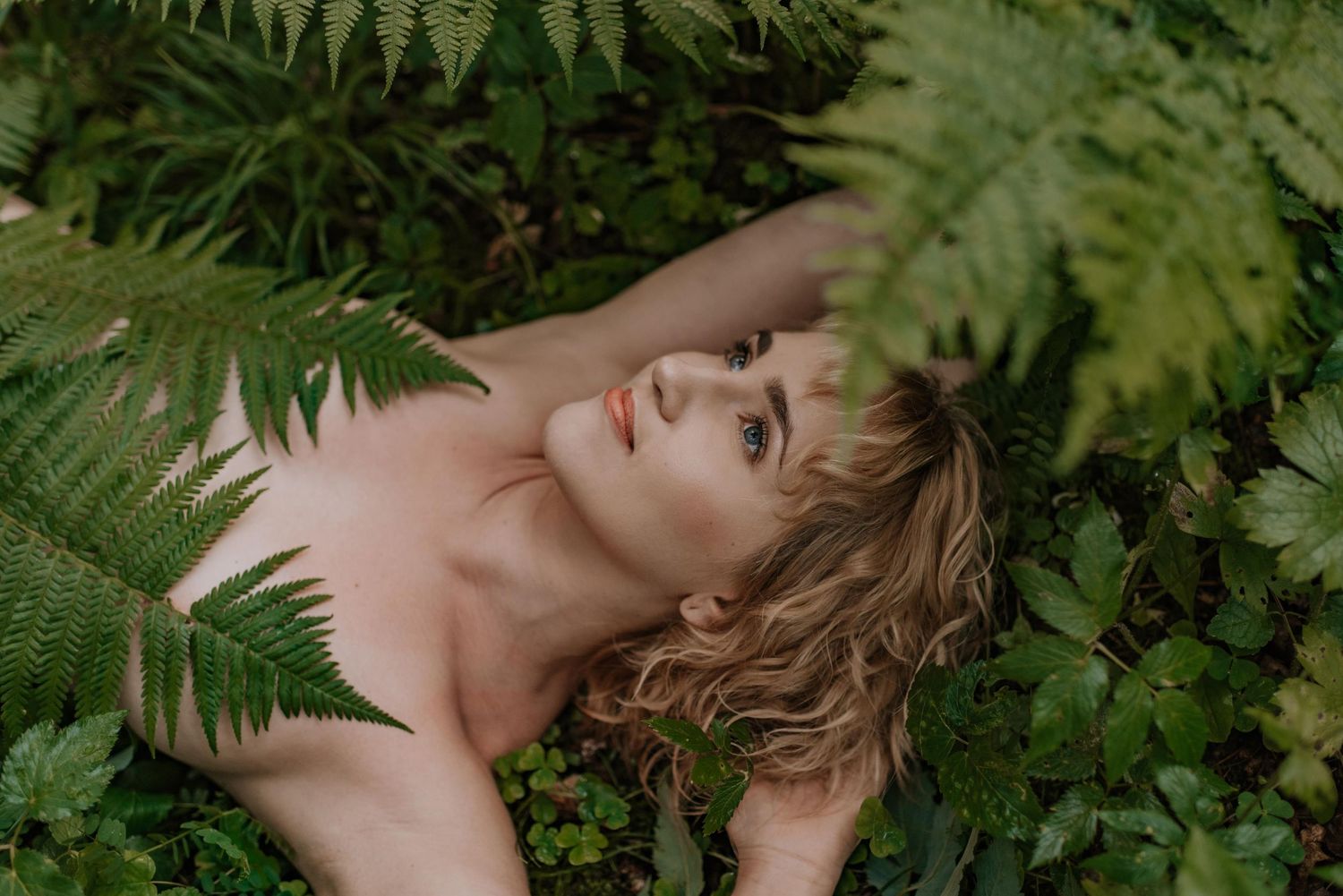 A person lying peacefully among green ferns and foliage in a natural outdoor setting.