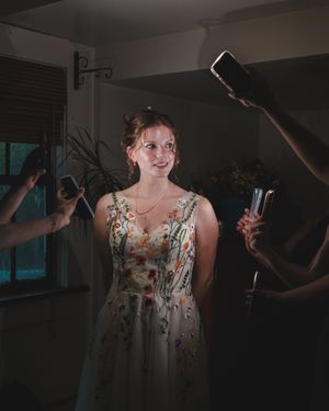 A woman in a floral dress stands in a dimly lit room while someone holds up a light for dramatic effect.