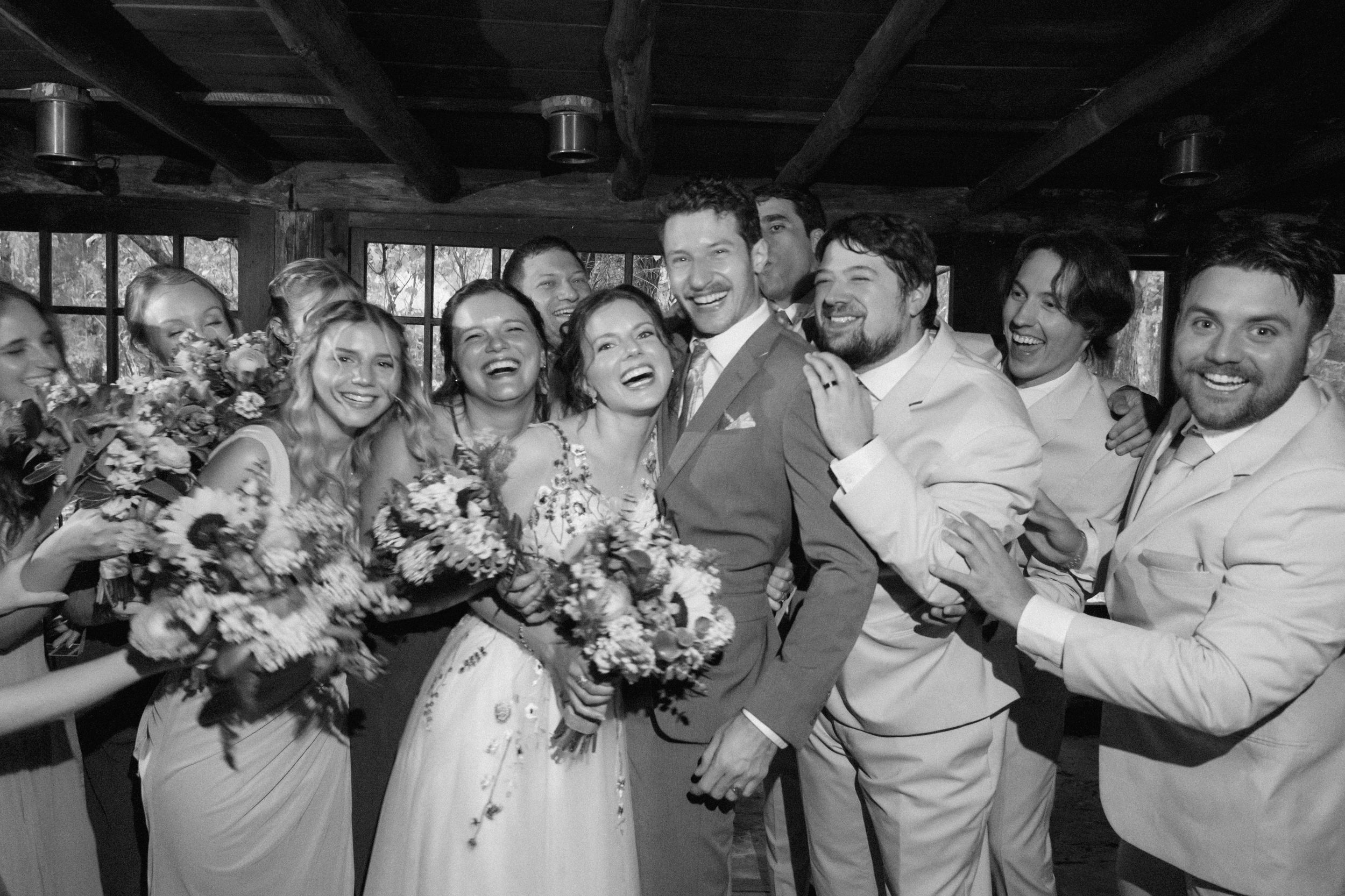 A black and white photo of a wedding party posing together with bouquets and laughing joyfully at an indoor venue.