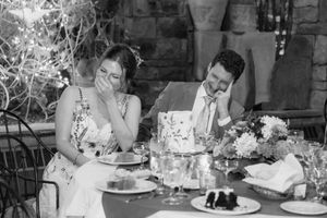A black and white photo of a couple sharing a laugh at an elegant outdoor dining table with floral centerpieces.