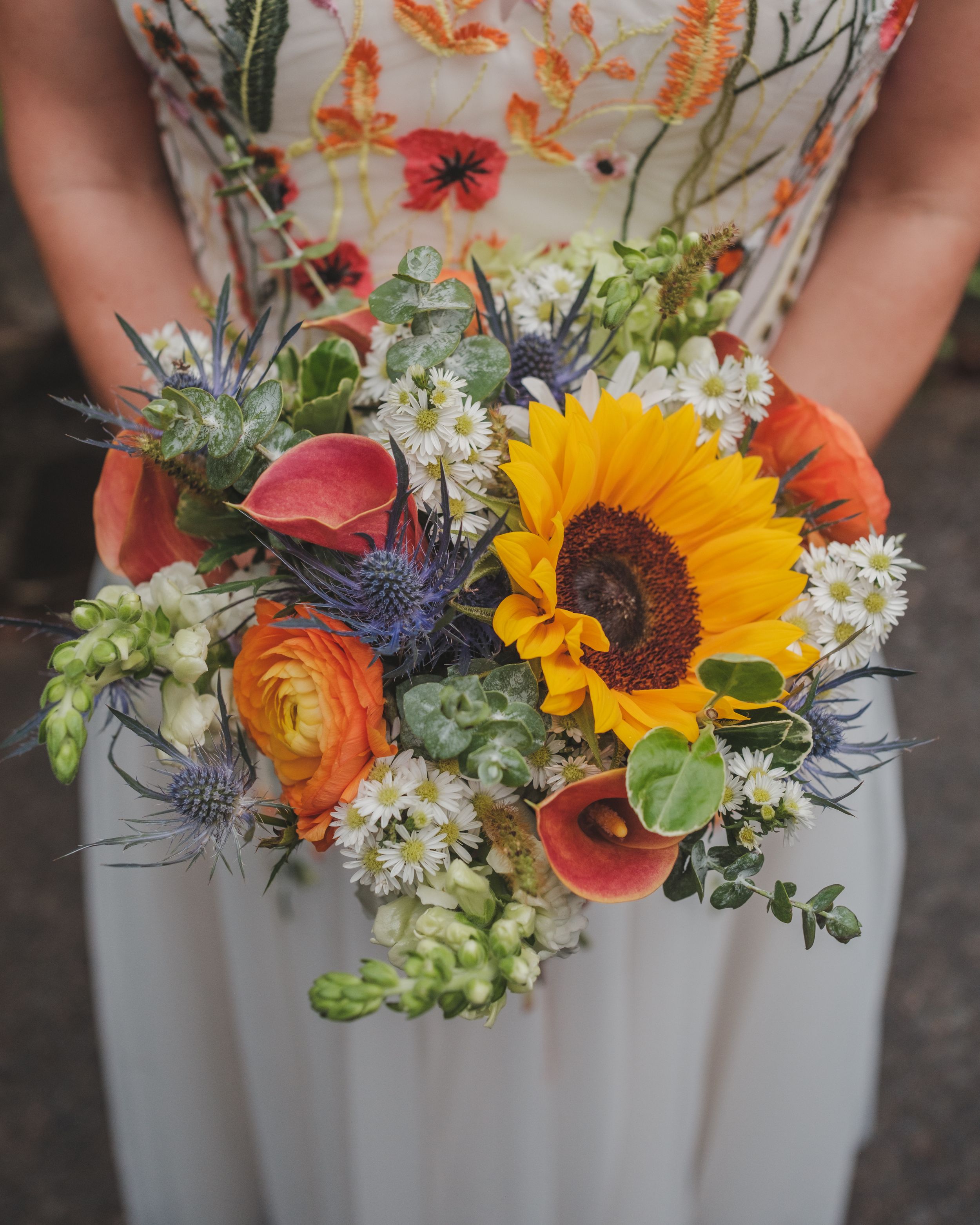 A vibrant bouquet featuring a sunflower, ranunculus, and wildflowers held against a floral patterned dress.