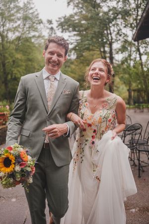 A joyful couple in wedding attire walk together outdoors, with the bride holding a sunflower bouquet.