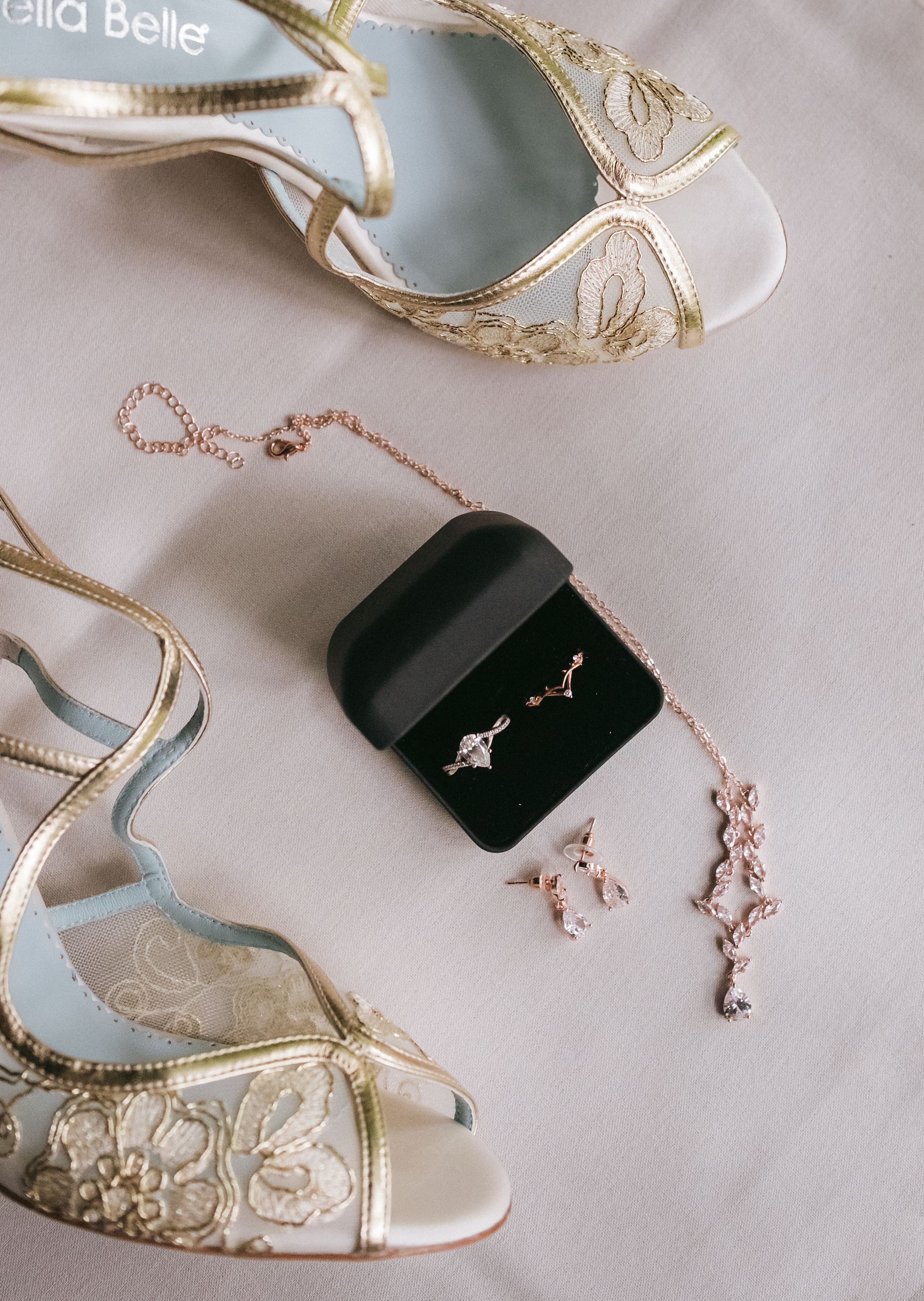 Elegant bridal accessories featuring white and gold lace shoes, a black ring box, and delicate rose gold jewelry on light fabric.