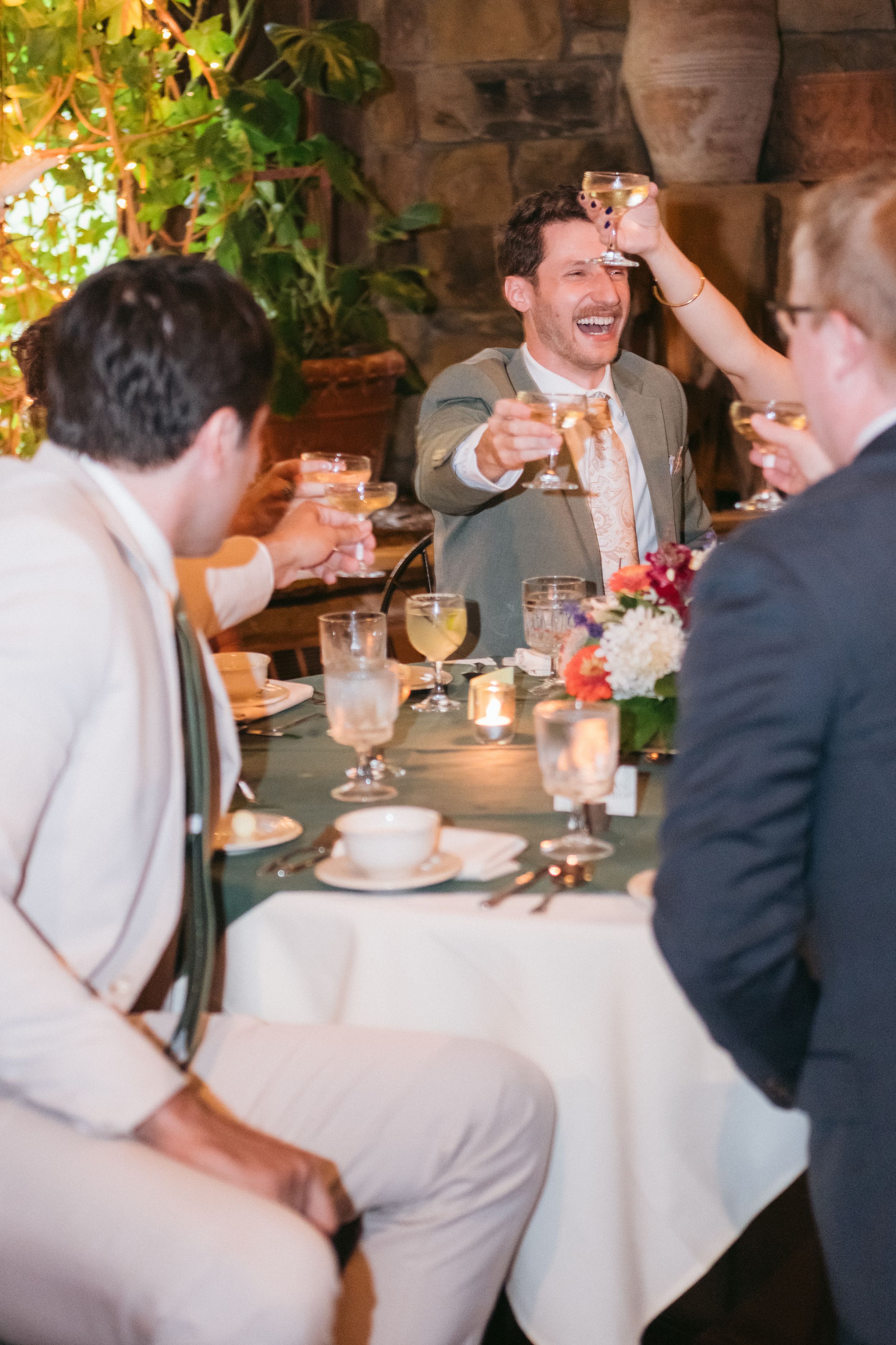 Wedding guests laugh and raise glasses during a reception dinner at an elegantly set table with floral centerpieces.