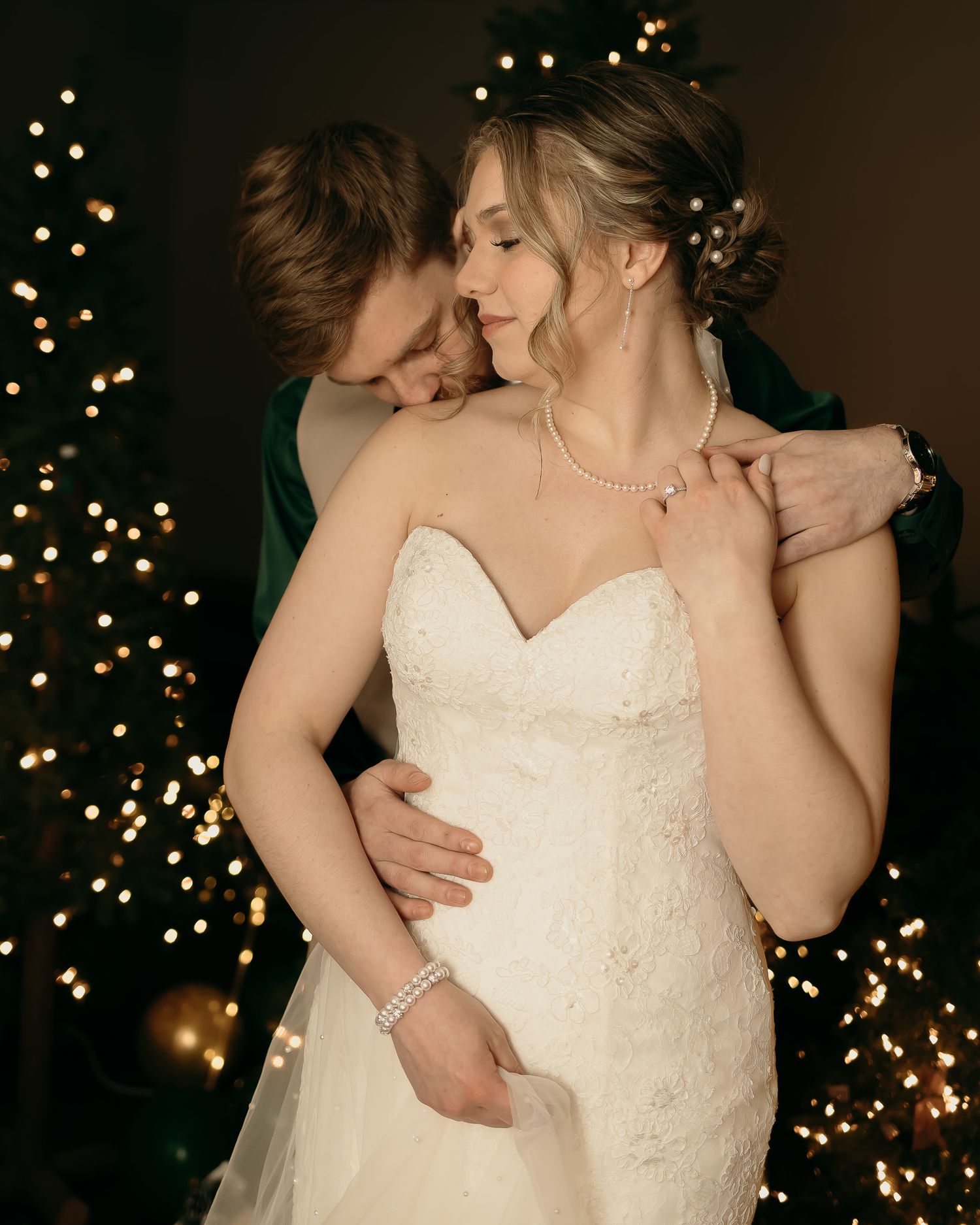Romantic wedding couple embraces in front of twinkling holiday lights, bride wearing strapless white lace gown.