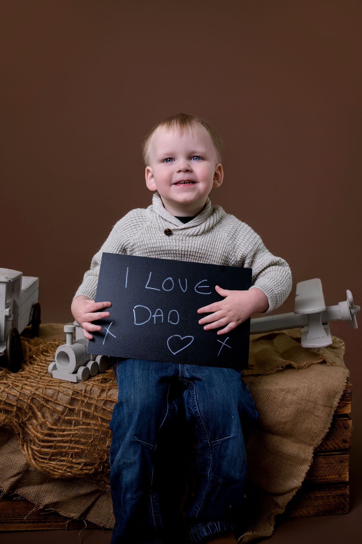Small child in a beige sweater holds a chalkboard sign with 'I Love Dad' written on it against brown background with decor.