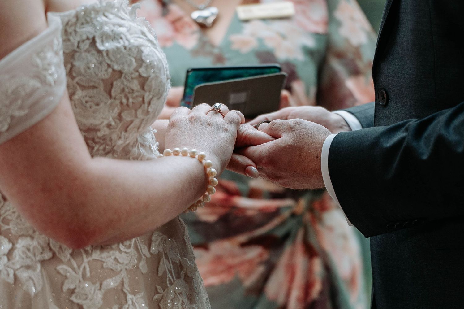 Close-up of hands exchanging rings during wedding ceremony with lace dress detail.