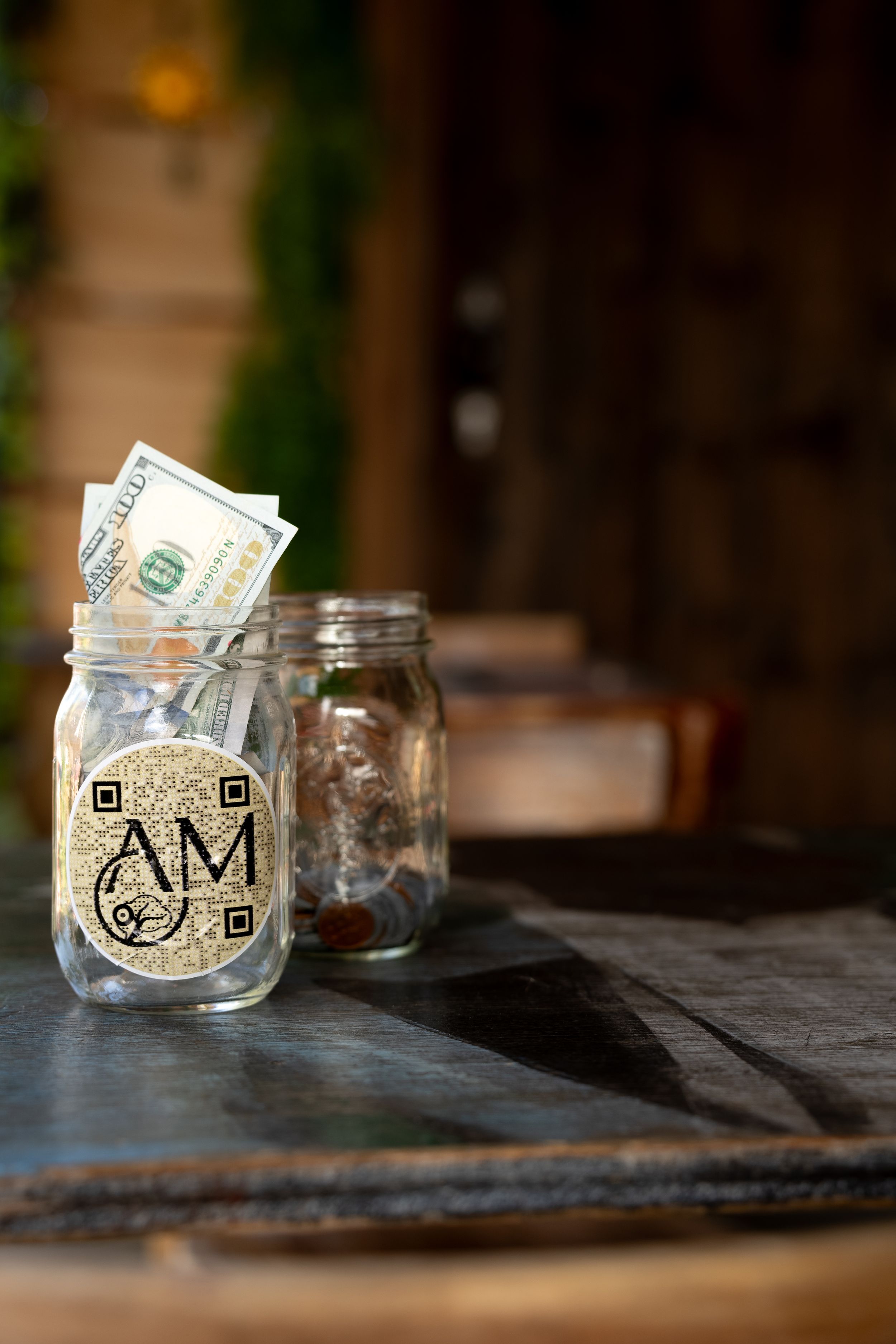 Clear mason jar with 'AM' logo and dollar bills sitting on dark wooden table with blurred background.